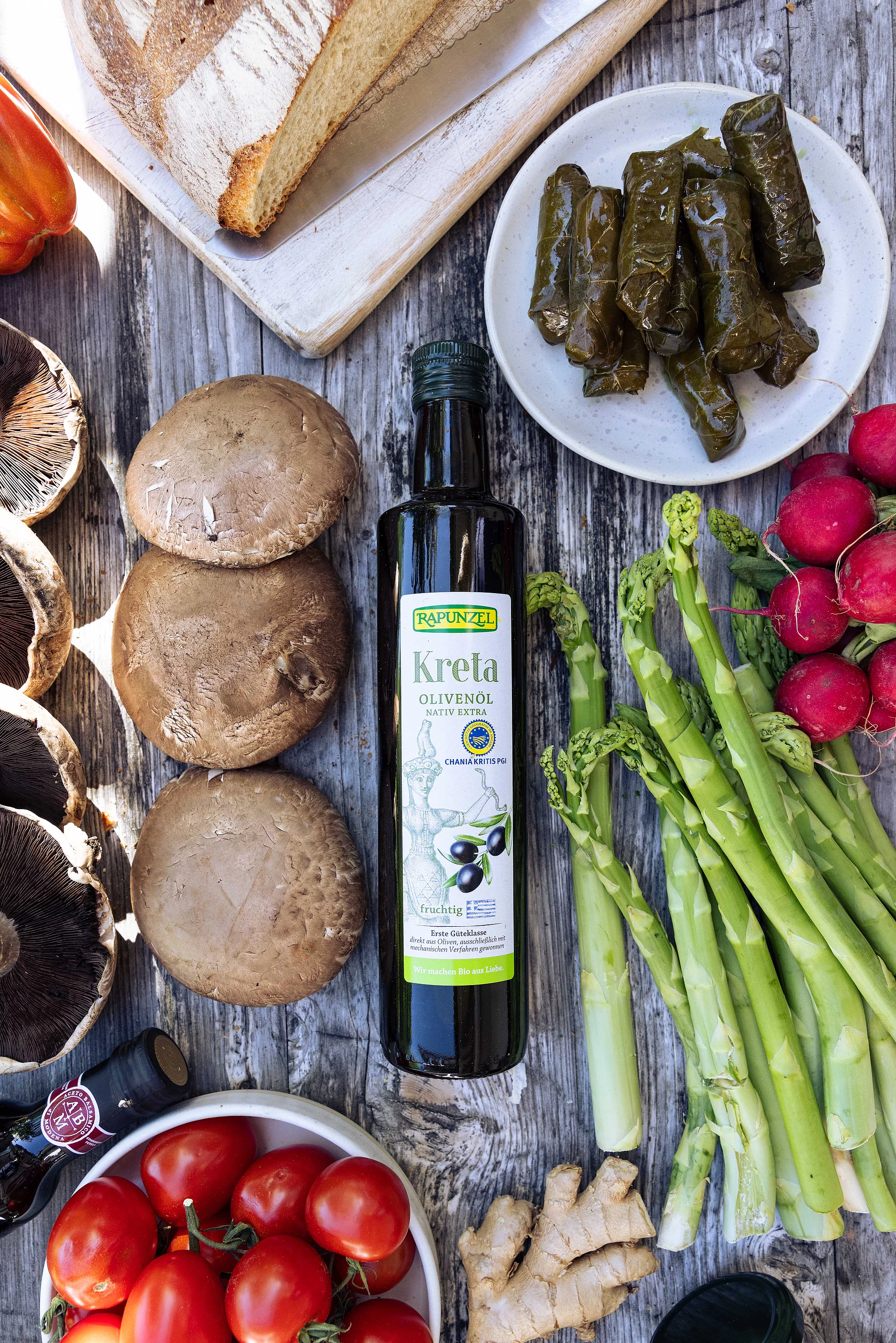 Flasche Rapunzel Olivenöl aus Kreta auf Tisch mit grünem Spargel, Pilzen und Weinblättern – vielseitig für mediterrane Gerichte.