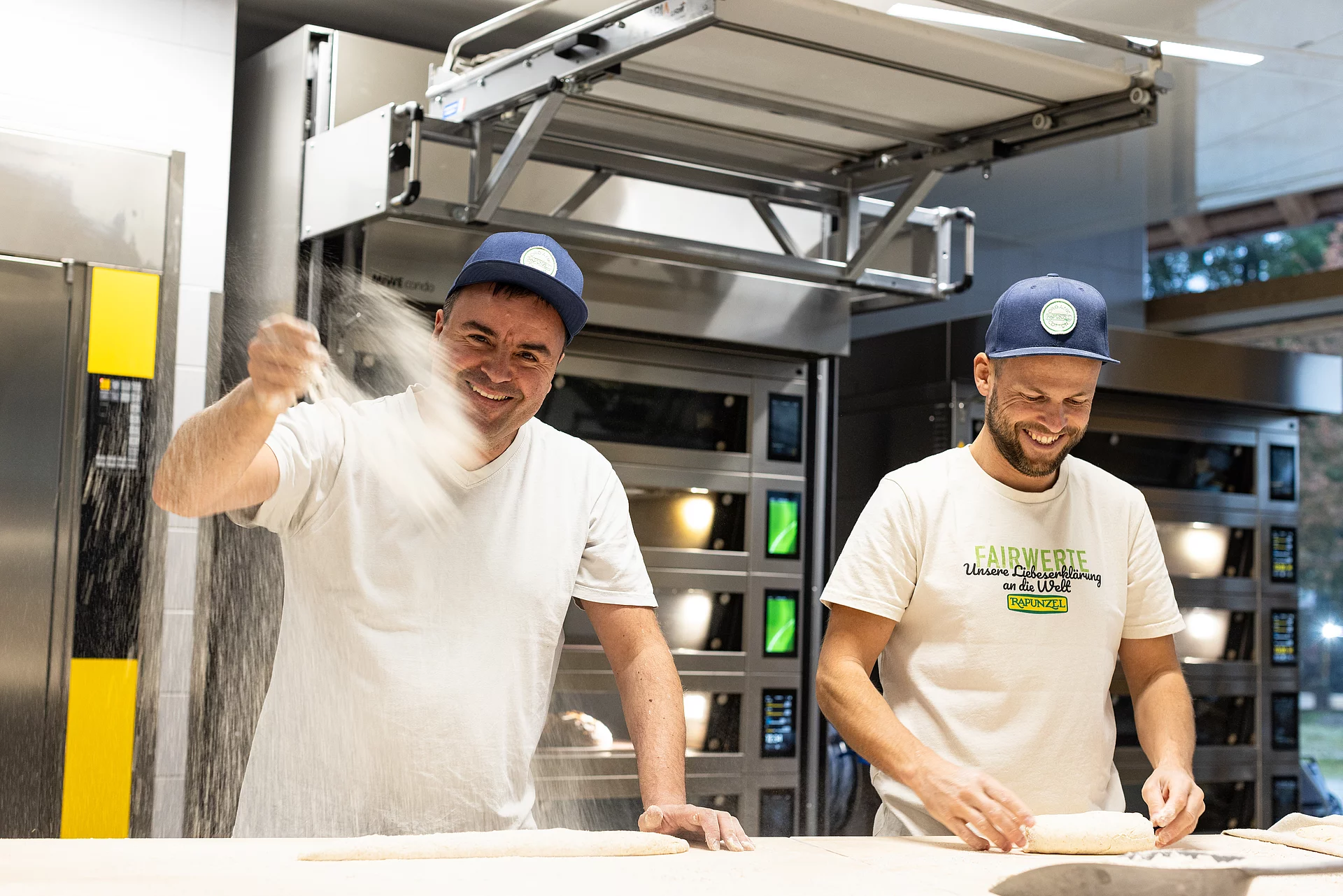 Zwei Bäcker am Backtisch in der Rapunzel Bäckerei bei der Arbeit, einer wirft Mehl in die Luft, beide lachen.
