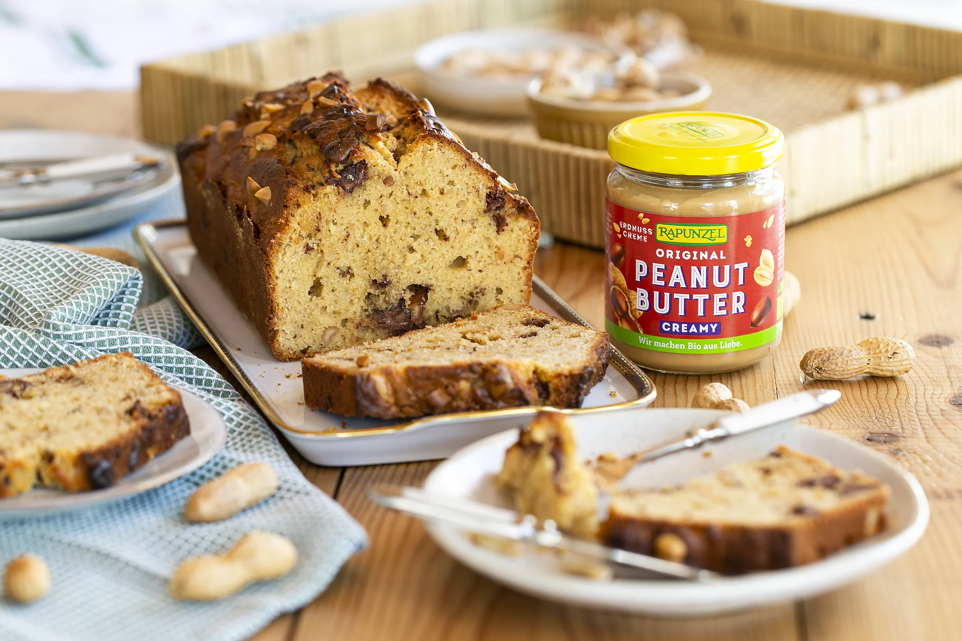 Ein Glas Peanutbutter steht auf einem Holztisch neben einem Kuchen. Einige Erdnüsse liegen daneben und ergänzen die natürliche Bildszene.
