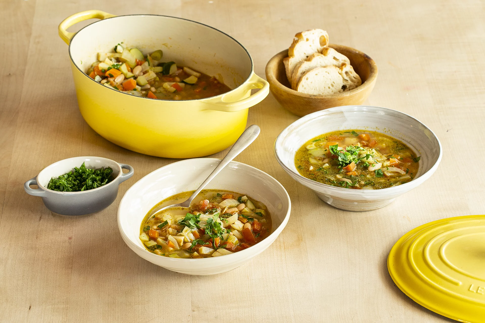 Zwei Teller mit Minestrone neben einem gelben Kochtopf und Schüsseln mit Brot und Kräutern.