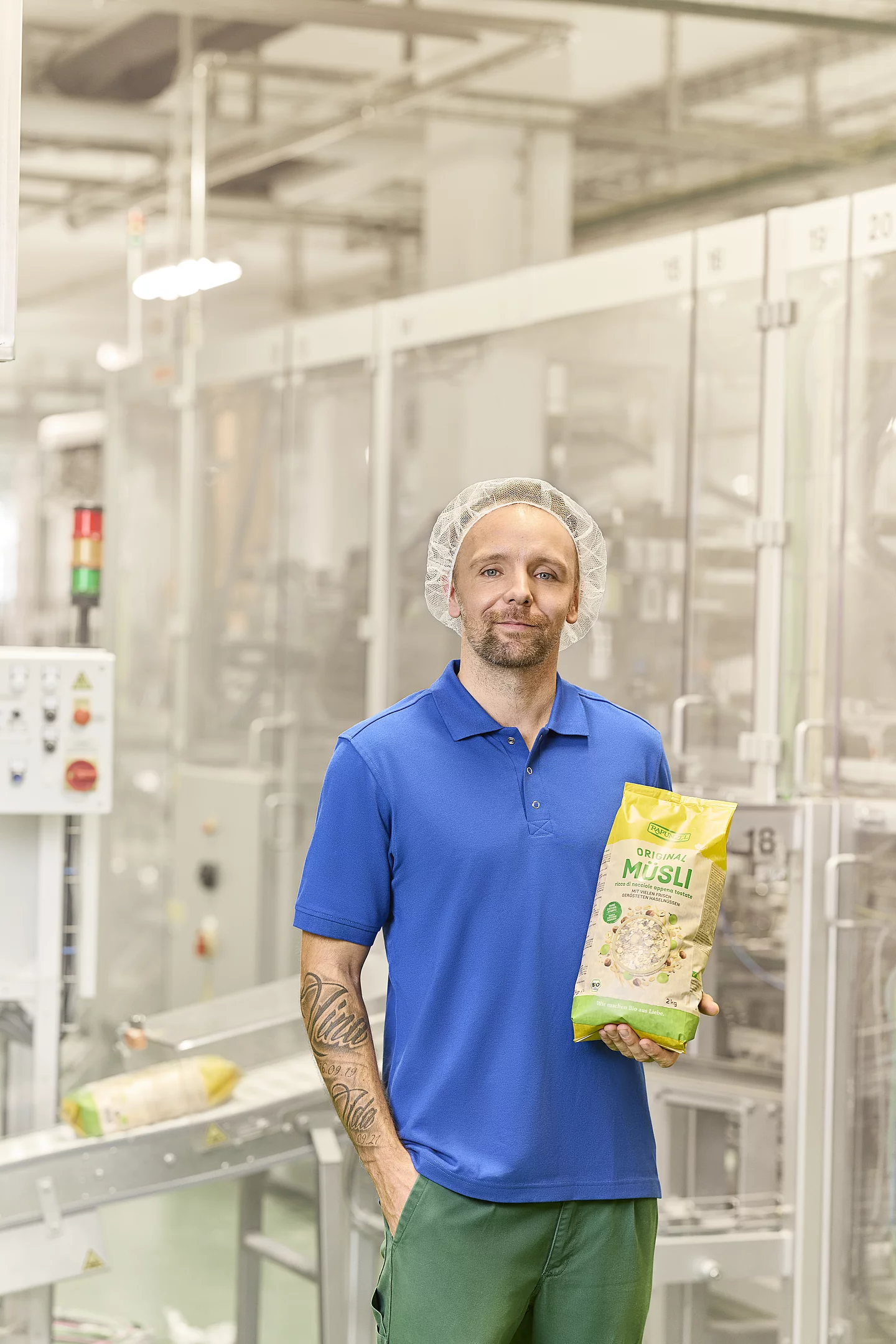 Eigene Herstellung: Müsli und Porridge Mitarbeiter aus der Produktion steht neben der Abpackmaschine und präsentiert fertig abgepacktes Müsli.
