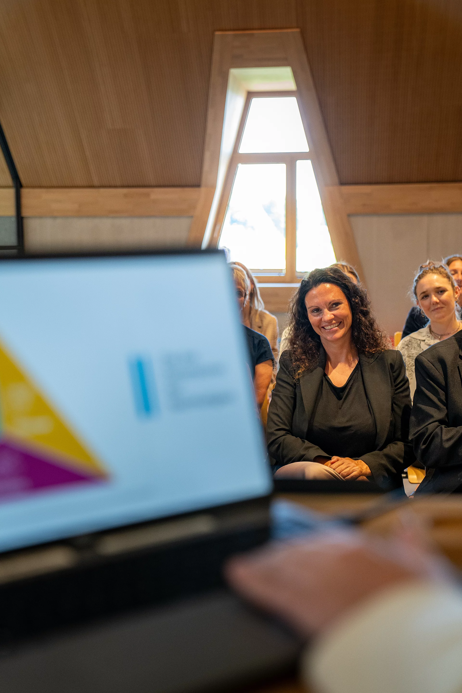 Im Vordergrund ein Laptop, im Hintergrund eine Frau, die interessiert zuhört.