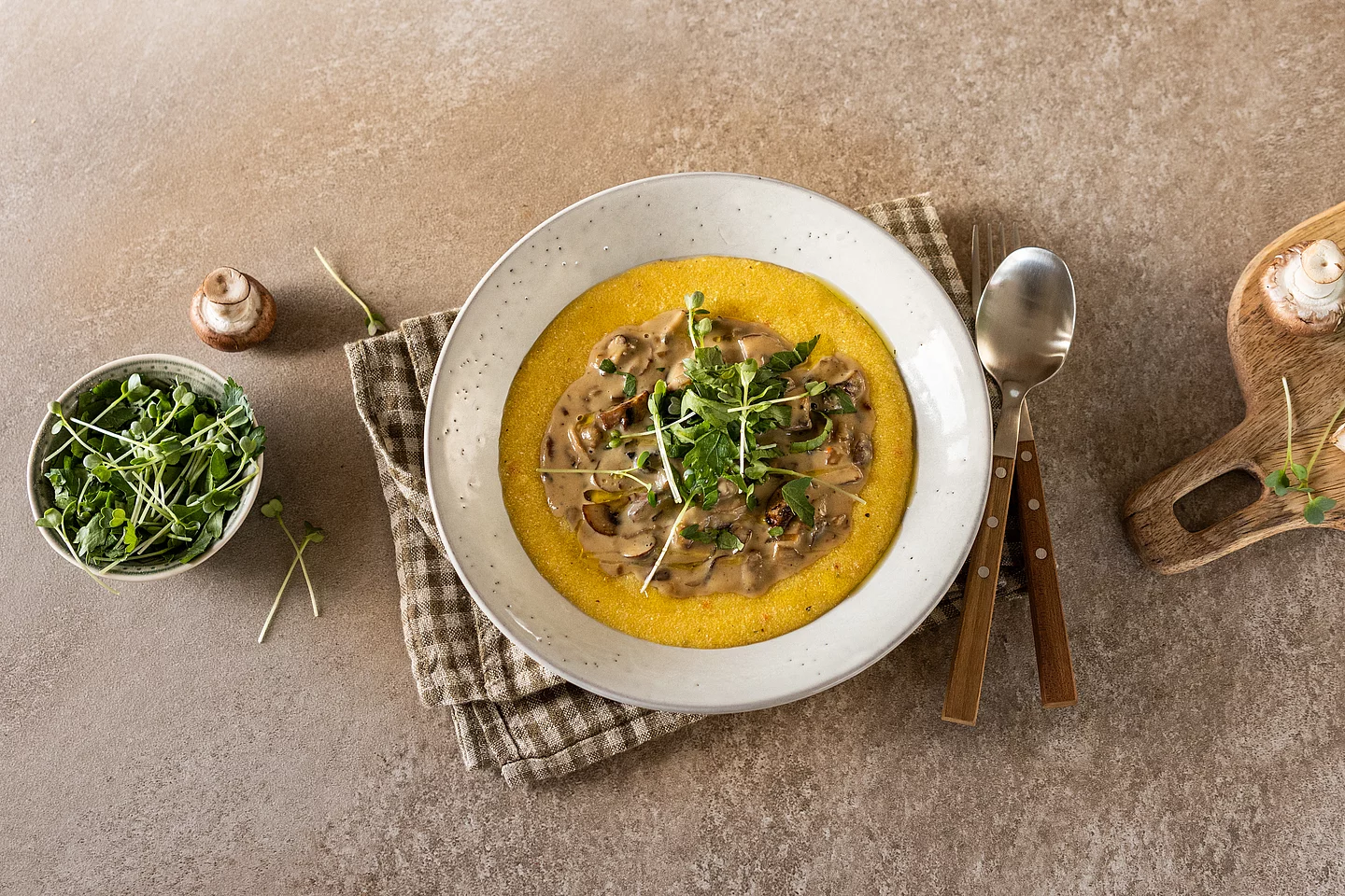 Eine Draufsicht auf weißen Teller mit leuchtend gelber, cremiger Polenta, in deren Mitte ein dunkles Pilz-Ragout in einer Sahnesauce angerichtet ist. Garniert mit frischen grünen Sprossen. Der Teller steht auf einer braun-weiß karierten Stoffserviette auf einer rustikalen Steinfläche. Daneben liegen ein Löffel und eine Gabel mit Holzgriffen sowie ein kleines Schälchen mit weiteren Sprossen und frischen Champignons.